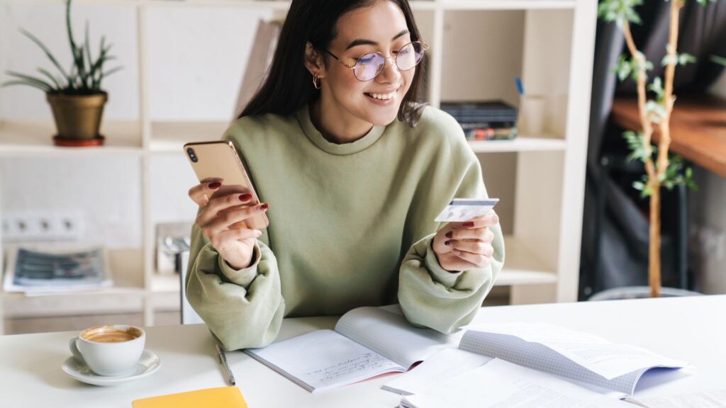 Young woman holding phone in one hand and credit card in the other. Notebooks are spread out on the table.